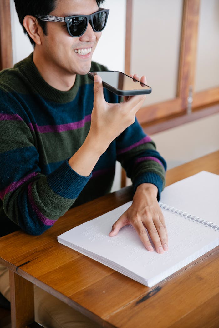 Smiling man uses smartphone for communication while reading Braille indoors.